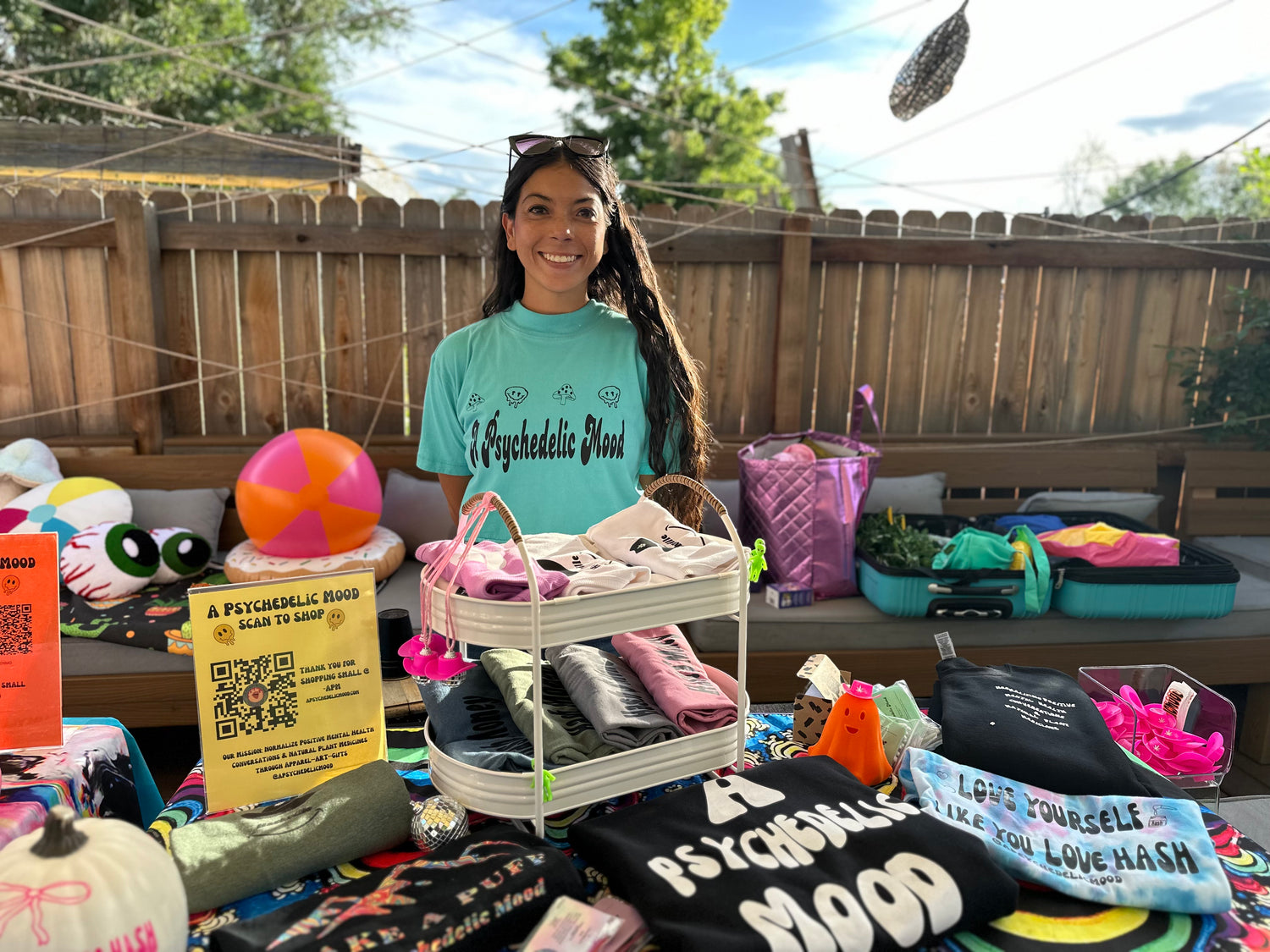 Person standing behind a table with various items including t-shirts and bags in an outdoor setting.
